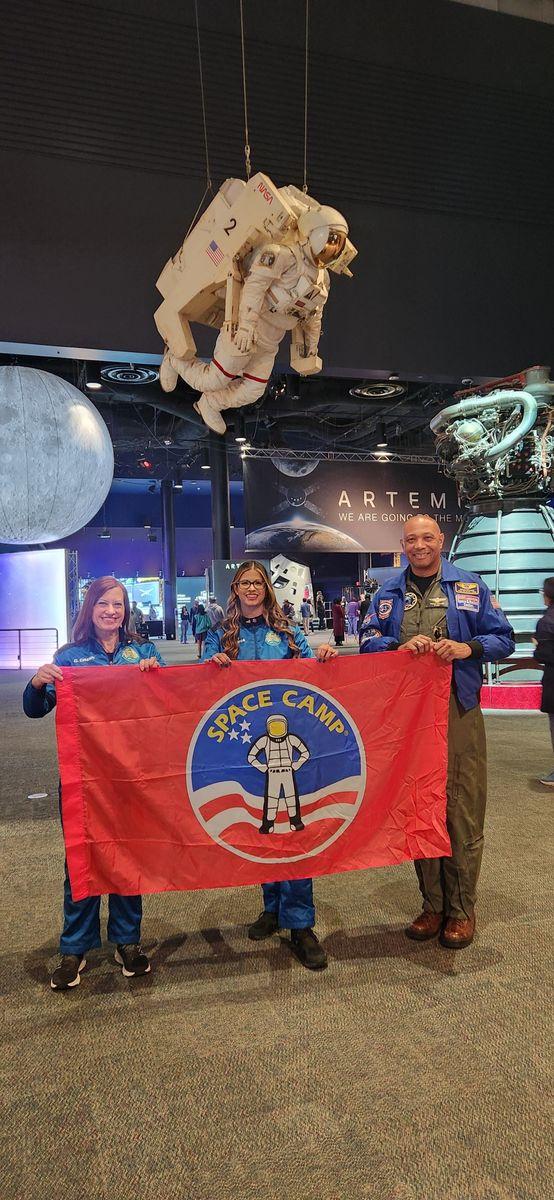 Dr. Gretchen Green and Diallo Wallace holding a Space Camp flag at the U.S. Space and Rocket Center with a spacesuit display and Artemis exhibit in the background